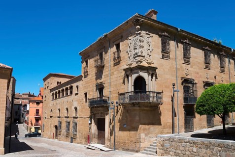 Historic sandstone city building with balcony and coat of arms under a clear sky, surrounded by