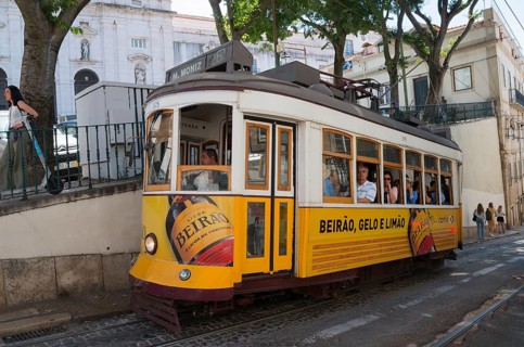 A yellow tram with Beirão advertising runs through a narrow city street, people on board, tram,