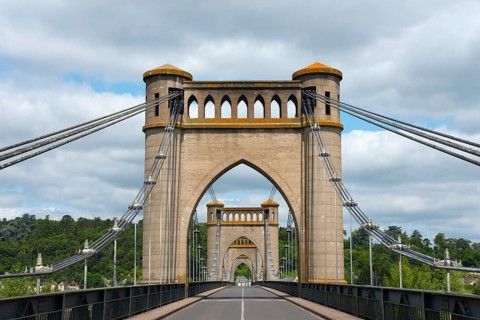 Historic bridge with distinctive arches and cables, surrounded by green landscape and a cloudy sky,