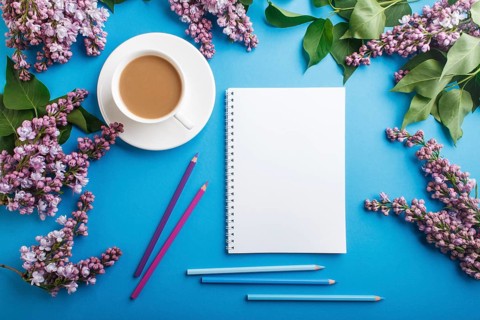 Purple lilac flowers and a cup of coffee with notebook and colored pencils on pastel blue