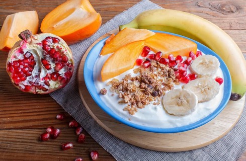 A blue plate with greek yogurt granola, persimmon, banana, pomegranate brown wooden background.