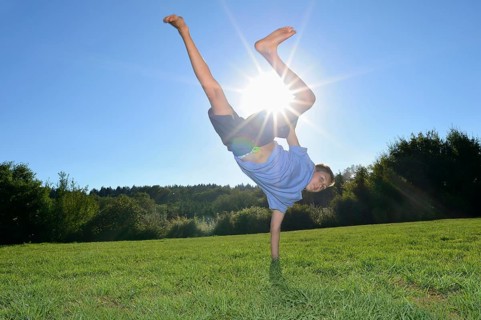 A boy doing a one-handed handstand on a grassy field with the sun shining brightly in the clear