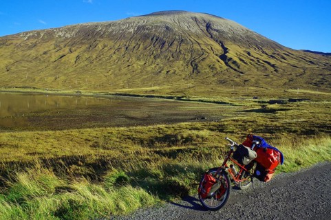 A loaded bicycle stands at the roadside in front of an impressive mountain landscape under a clear