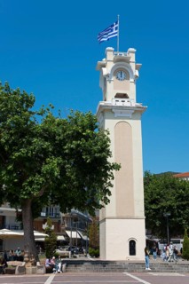 A tower clock with the Greek flag surrounded by trees and people in a relaxed atmosphere, Xanthi,