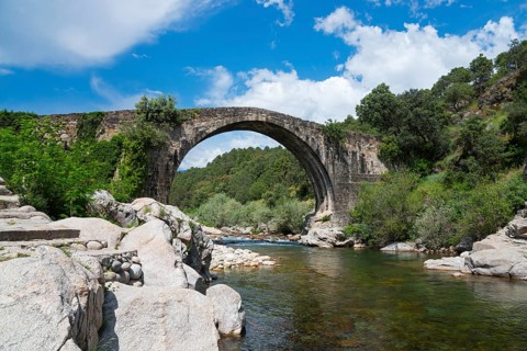 Steinbrücke über einem ruhigen Fluss mit felsiger Uferlandschaft und dichtem Grün unter einem blau