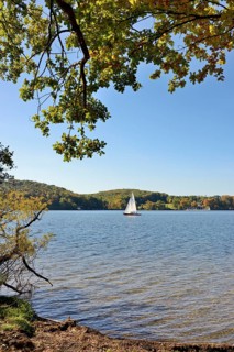 Deciduous trees with autumn leaves on the lakeshore, sailing boat on the Möhne dam, blue sky, North