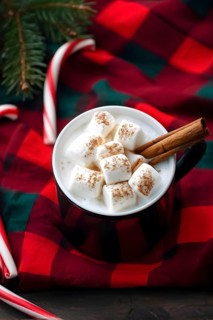 A close-up of a steaming cup of hot cocoa topped with marshmallows, surrounded by cinnamon sticks,