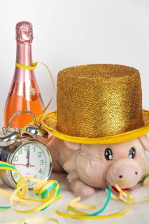 Plush pig wears golden hat next to alarm clock and champagne bottle, New Year's Eve