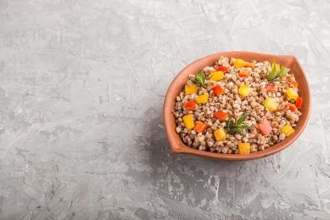 Buckwheat porridge with vegetables in clay bowl on a gray concrete background. Side view, copy