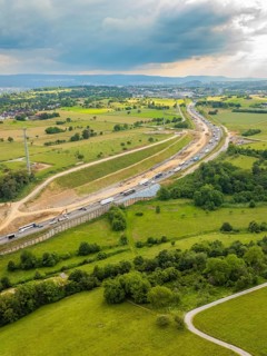 View of a large construction site next to a road through green fields and under a cloudy sky, A8