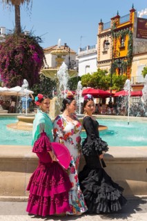Three woman in colourful flamenco dresses, with flowers in their hair, stand in front of a fountain