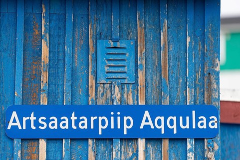 A blue sign inscribed with a Greenlandic street name on a weather-beaten wooden wall, remote Arctic
