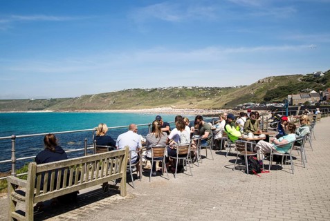 People sitting outdoors at cafe tables by the sea, Sennan Cove, Land's End, Cornwall, England, UK