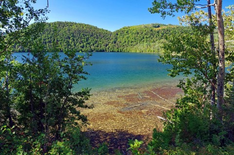 A clear lake seen from a wooded shore surrounded by green mountains under a blue sky, Gaspesie,