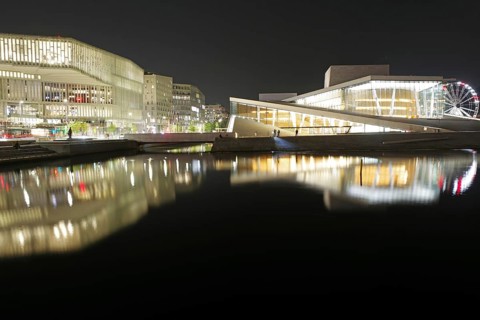 Modern buildings and opera house with glowing facades reflected in calm waters at night, Oslo,