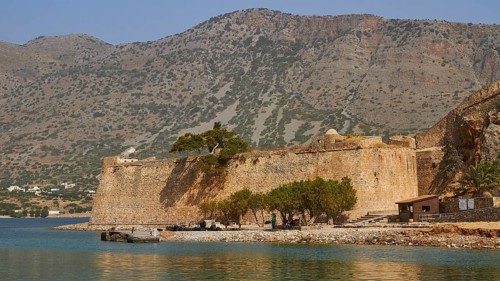 Old fortress on the coast, surrounded by trees and water, with hilly landscape in the background,