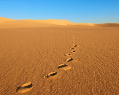Footprints run through a quiet, sunlit desert with large sand dunes and a clear blue sky, Matruh,