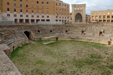 An ancient Roman amphitheatre made of stone with grass inside, surrounded by historic buildings,