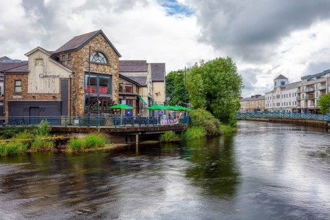 Historic buildings and cafés along a river with green vegetation and clouds, Sligo
