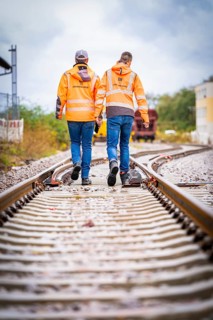 Two men in orange walking hand in hand on railway tracks in an unclear direction, track