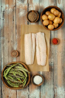 Two fresh cod fish fillets on parchment paper surrounded by various spices, green beans, and small