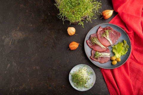Slices of smoked salted meat with cilantro microgreen on black concrete background and red textile.