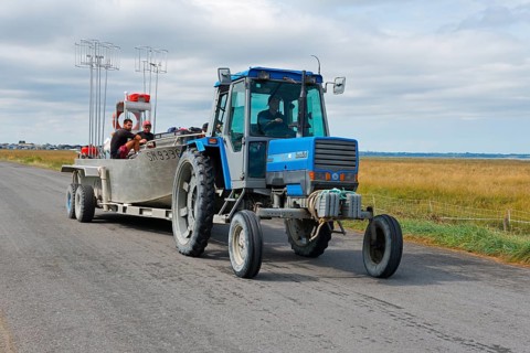 A blue tractor on a country road with people in the trailer, rural surroundings, tractor with boat