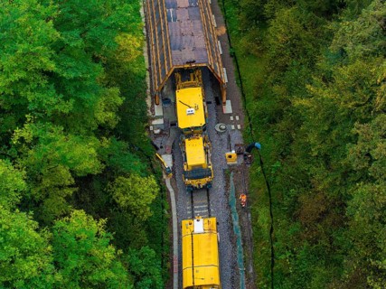 Yellow train exits a tunnel surrounded by forest while workers stand on the tracks, tamping