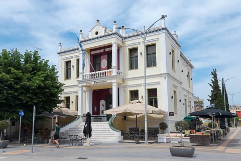 A magnificent neoclassical building with cafés in front and a clear blue sky, Komotinaion Club,
