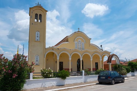 Church with yellow facade and bell tower under a blue sky, surrounded by flowers and a parked car,