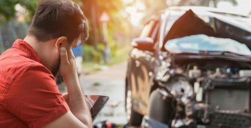 A man sits on the ground next to a car that has been in an accident. He is looking at the car with