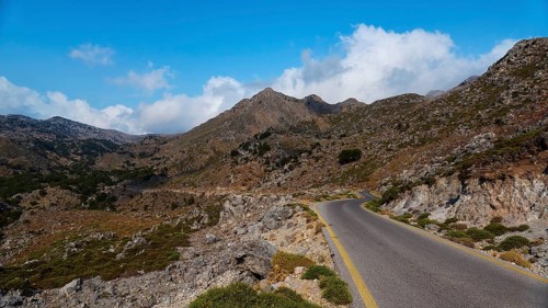 Asphalt road winds through a rocky mountain landscape under a blue, slightly cloudy sky,