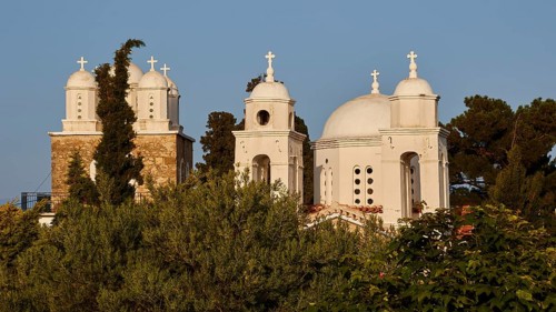 White church with three domes and bell towers, surrounded by greenery under a clear sky, Koroni,