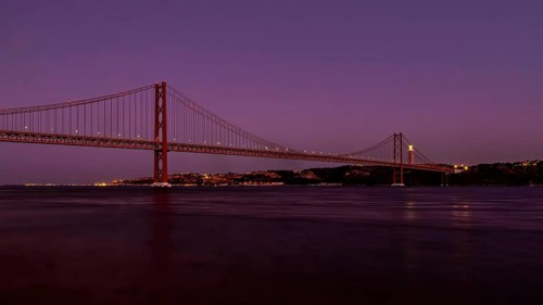 Twilight descends on a calm river with a suspension bridge standing majestically against a purple