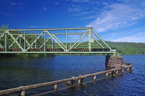 A green metal bridge crosses a river in a natural setting with trees and blue sky, railway bridge,