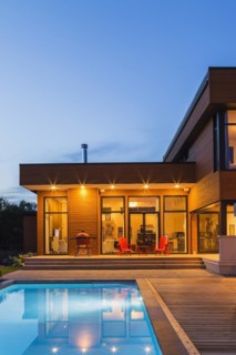 Modern cube style home with stained horizontal wood cladding and in-ground swimming pool at dusk,