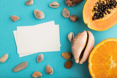 White paper business card with ripe cut papaya, orange, seashells on blue pastel pastel background.
