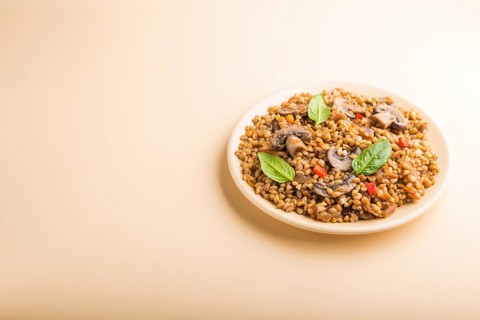 Spelt (dinkel wheat) porridge with vegetables and mushrooms on ceramic plate on orange background.
