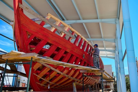 A partially built red wooden ship in a covered shipyard with a labourer working on it, Patmos
