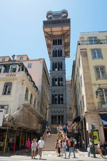 Tower-like lift between old buildings, people walking up the stairs, clear sky, sunny weather,