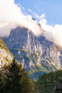 Impressive mountains shrouded in clouds, surrounded by autumn-coloured trees under a clear blue