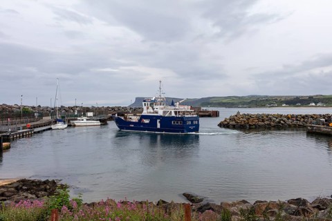 A blue ship sails in a cloudy harbour with rocky shores and blue water, Ballycastle