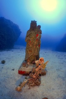A statue of a mother and child, Mary and Jesus, stands on a sandy seabed underwater. Dive site