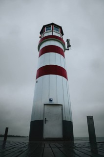 A red and white lighthouse stands on a wooden plank under a grey sky, Podersdorf, Burgenland,