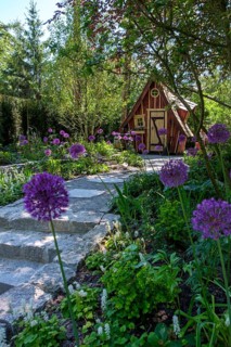 View of a garden with a wooden hut and a stone path, lined with purple flowers and surrounded by