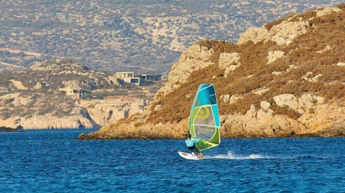 Person windsurfing along a rocky coast on blue water, windsurfer, Meltemi windsurfing spot, Devils