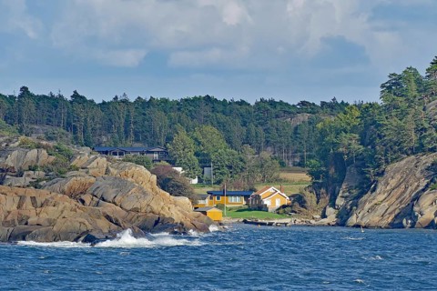 Coastal view with a wooded hill, rocks on the shore, several houses and waves under a cloudy sky,