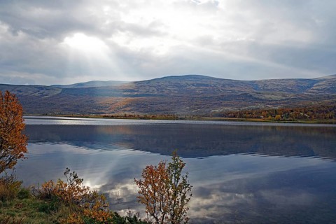 A lake with mountains in the background, rays of light penetrate the clouds, autumn leaves and