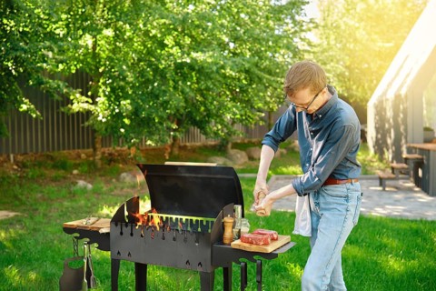 Middle-aged man sprinkles raw steak with sea salt using hand mill while cooking at backyard