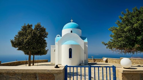 White-blue church behind a lattice gate with surrounding trees under a blue sky, Profitis Ilias